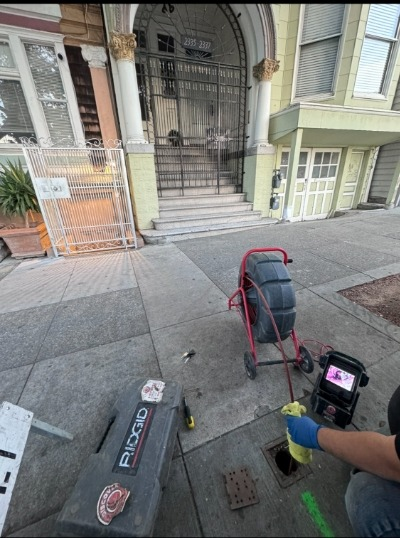 A technician from DiscoverPlumbing&Rooter in San Francisco uses a camera to inspect a sewer line, with various tools visible on the sidewalk.